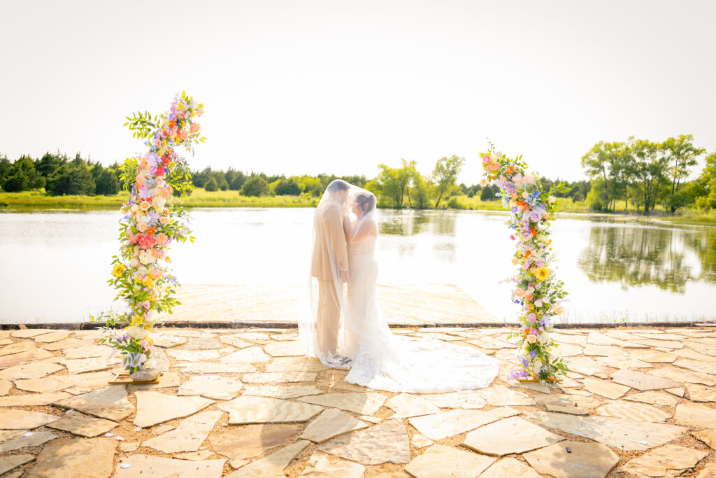 Bride and groom standing together under the Bride's veil. The couple are standing between two tall floral arrangements with a water front behind them. An Austin wedding photo and video team covered this wedding day.