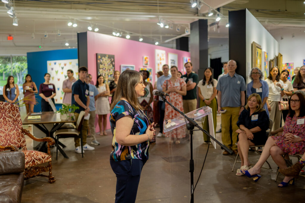 A woman speaking to a crowd of listeners in an art gallery for a fundraiser event. Photo was taken by a local Austin event coverage team.