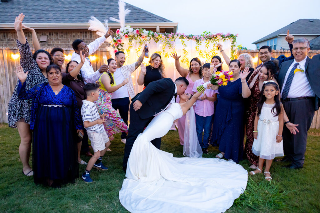 Bride and groom in the center of the photo. Bride is being dipped by the groom and they're kissing. They are surrounded by family and friends smiling and cheering at the couple. This photo was taken in by a Texas bilingual wedding photographer.